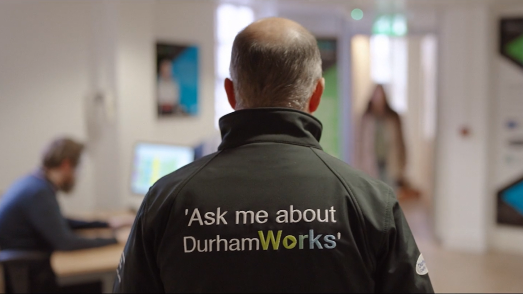Man standing with back to camera and words "Ask me about DurhamWorks" printed on the back of his jacket