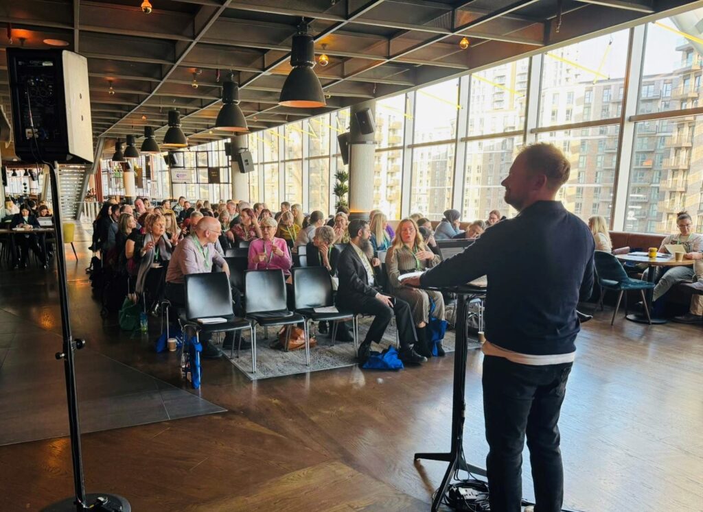 Ramsay Taylor stands behind a podium delivering a workshop to a large group of people in a large open room.